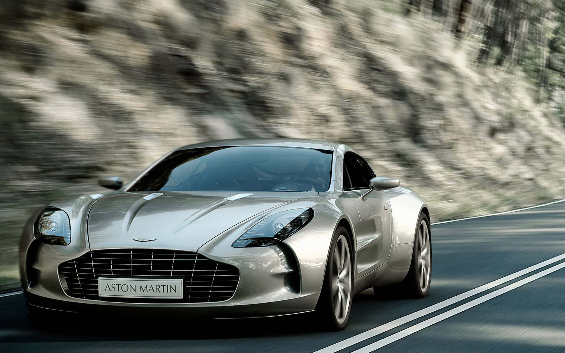 奧斯頓馬丁 桌布 壁紙 — 一輛銀色Aston Martin跑車沿著蜿蜒的山路疾馳，背景是模糊的岩石懸崖。