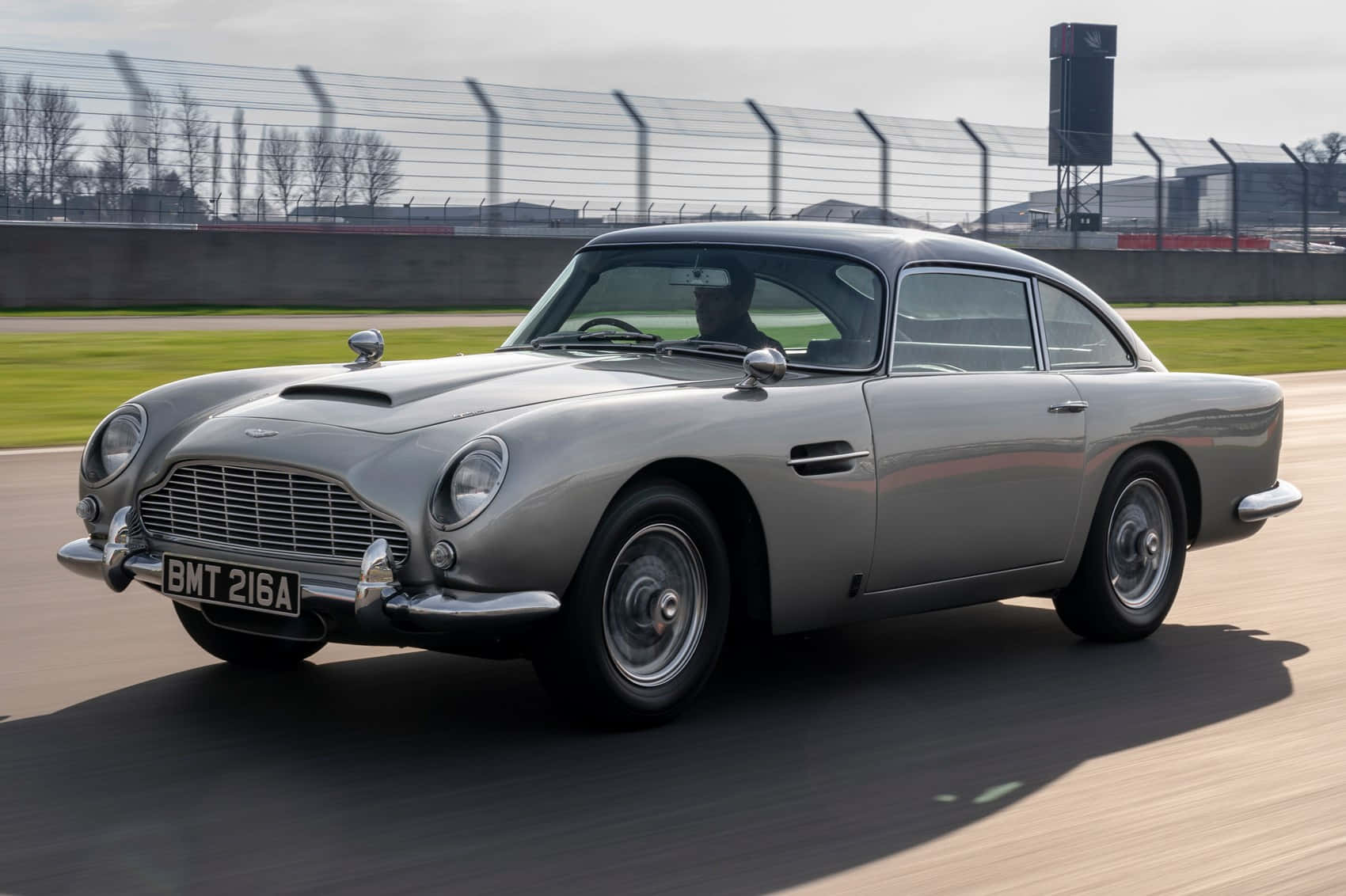 Aston Martin Db5 桌布 壁紙 — 一輛銀色Aston Martin DB5在賽道上疾馳，動作模糊，透過擋風玻璃可見駕駛員。