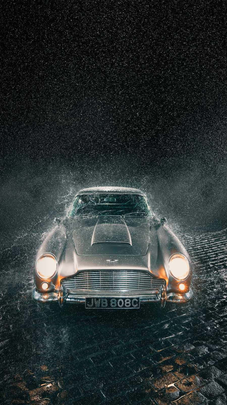 奧斯頓馬丁 桌布 壁紙 — 一輛銀色Aston Martin DB5從濕滑的鵝卵石中出現，閃爍的車頭燈刺穿黑暗的暴風雨之夜。