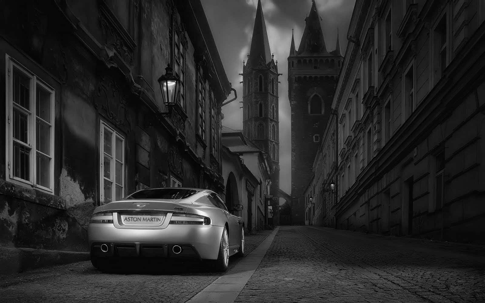 Cool Black And White 桌布 壁紙 — 一輛銀色Aston Martin停泊在鵝卵石街道上，下方是陰沉天空下高聳的哥德式塔樓。