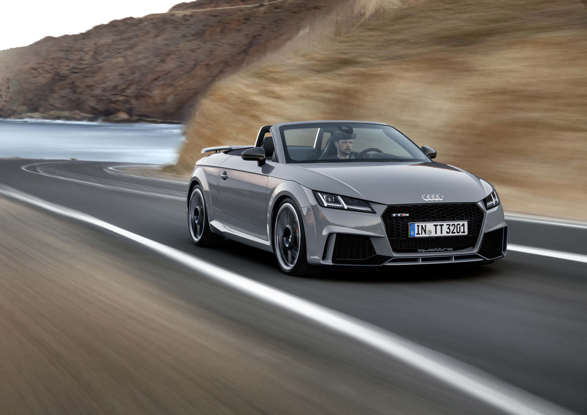 Audi Tt Rs 桌布 壁紙 — 一輛銀色Audi TT Roadster沿著海岸公路飛馳，背景是模糊的懸崖和移動的海洋。