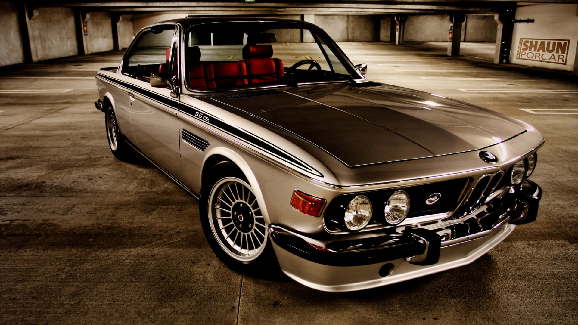 1920x1080 Bmw 桌布 壁紙 — 一輛銀色BMW 3.0 CSL配紅色內裝，停放在燈光昏暗、帶有暖色照明的混凝土車庫中。