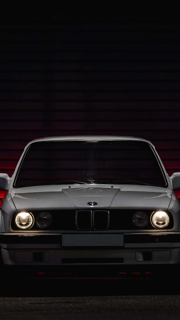 桌布 壁紙 — 一輛銀色BMW E30跑車，在昏暗光線下，亮著車頭燈，面向深紅色條板牆。