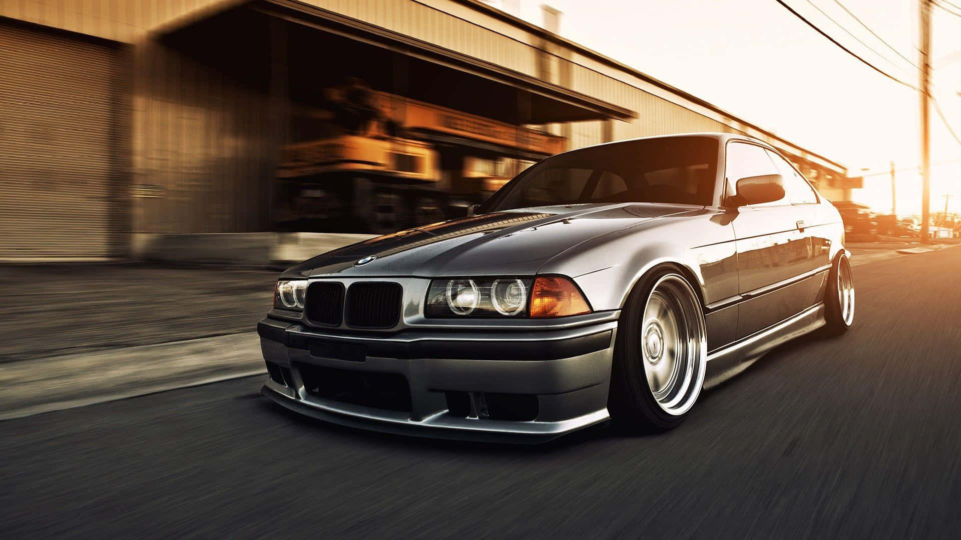 Tuned Car 桌布 壁紙 — 一輛銀色BMW E36雙門轎跑車在日落時分，伴隨動態模糊和溫暖的金色光線，在都市街道上疾馳。