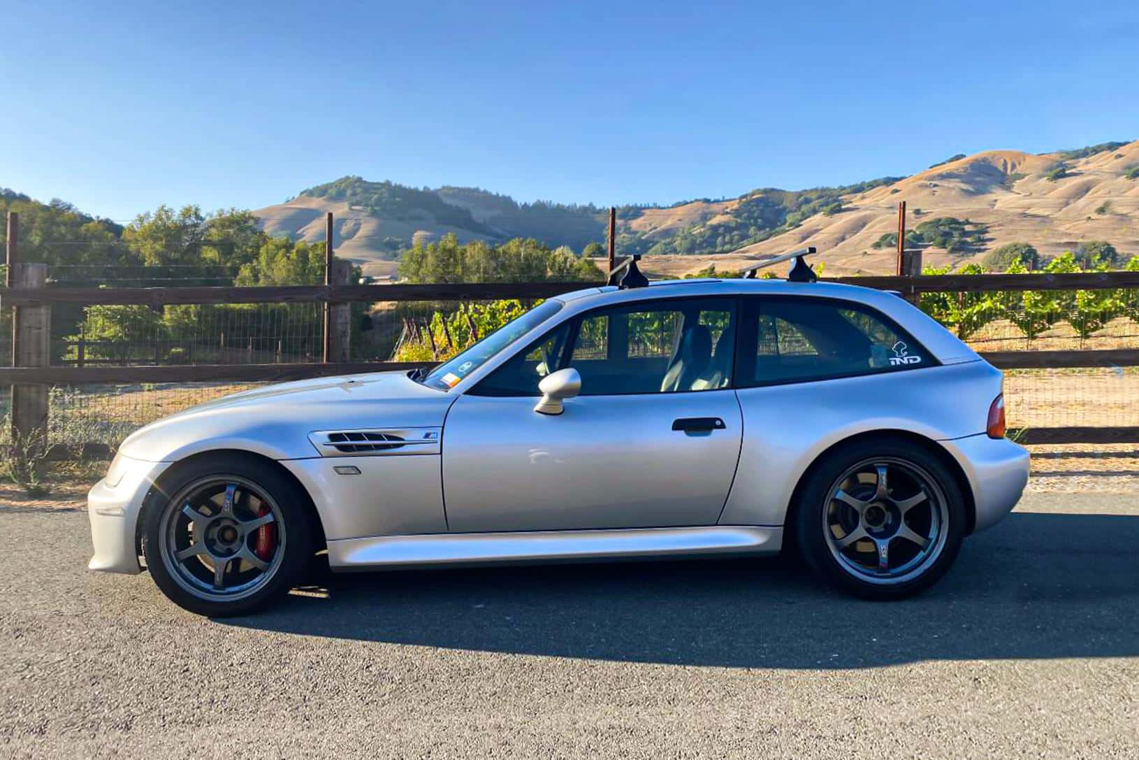 BMW 動圖 桌布 壁紙 — 一輛銀色BMW M3停泊在碎石上，背景是連綿起伏的山丘和葡萄園，天空清澈湛藍。