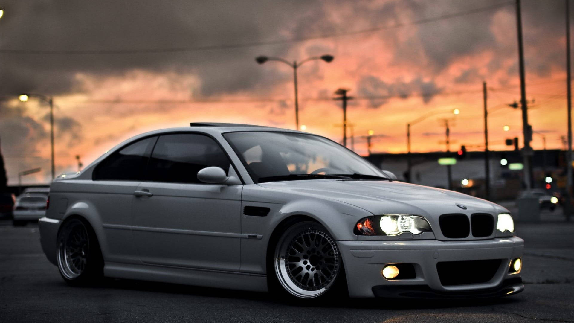 Lowrider 桌布 壁紙 — 一輛銀色BMW M3轎跑車在火紅的夕陽天空下閃耀，車頭燈發光，背景是城市街燈。