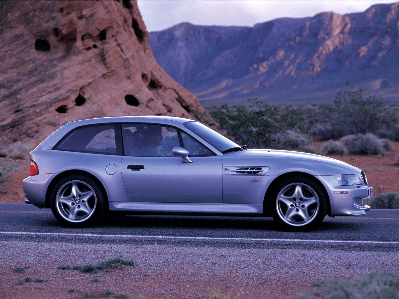Bmw Z3 桌布 壁紙 — 一輛銀色BMW M3運動旅行車停放在沙漠道路上，旁邊是紅色岩石地貌，天空昏暗。