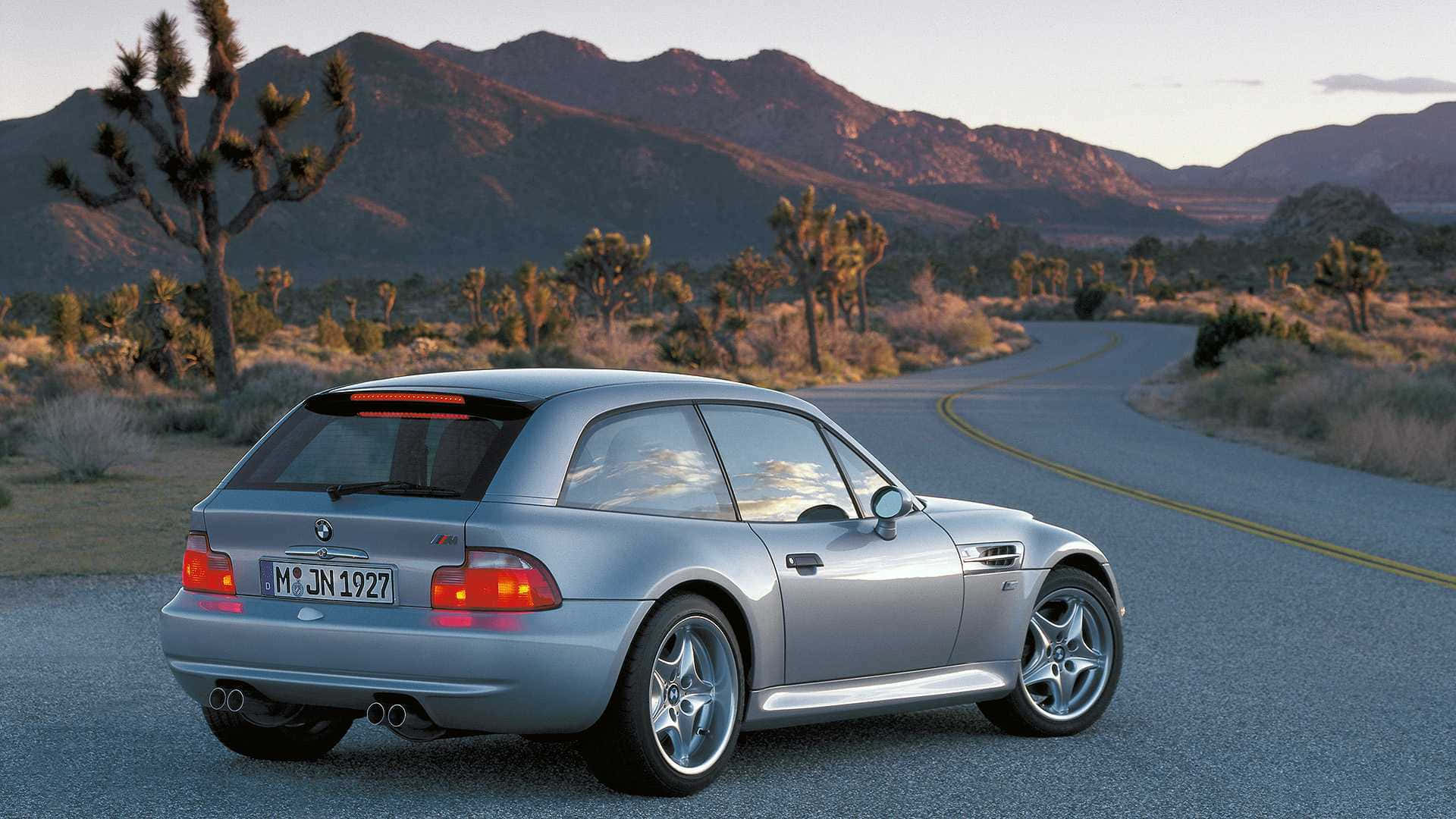 Bmw Z3 桌布 壁紙 — 一輛銀色BMW M3雙門跑車在蜿蜒的沙漠公路上行駛，兩旁有約書亞樹和山脈，天空呈現柔和的日落色彩。