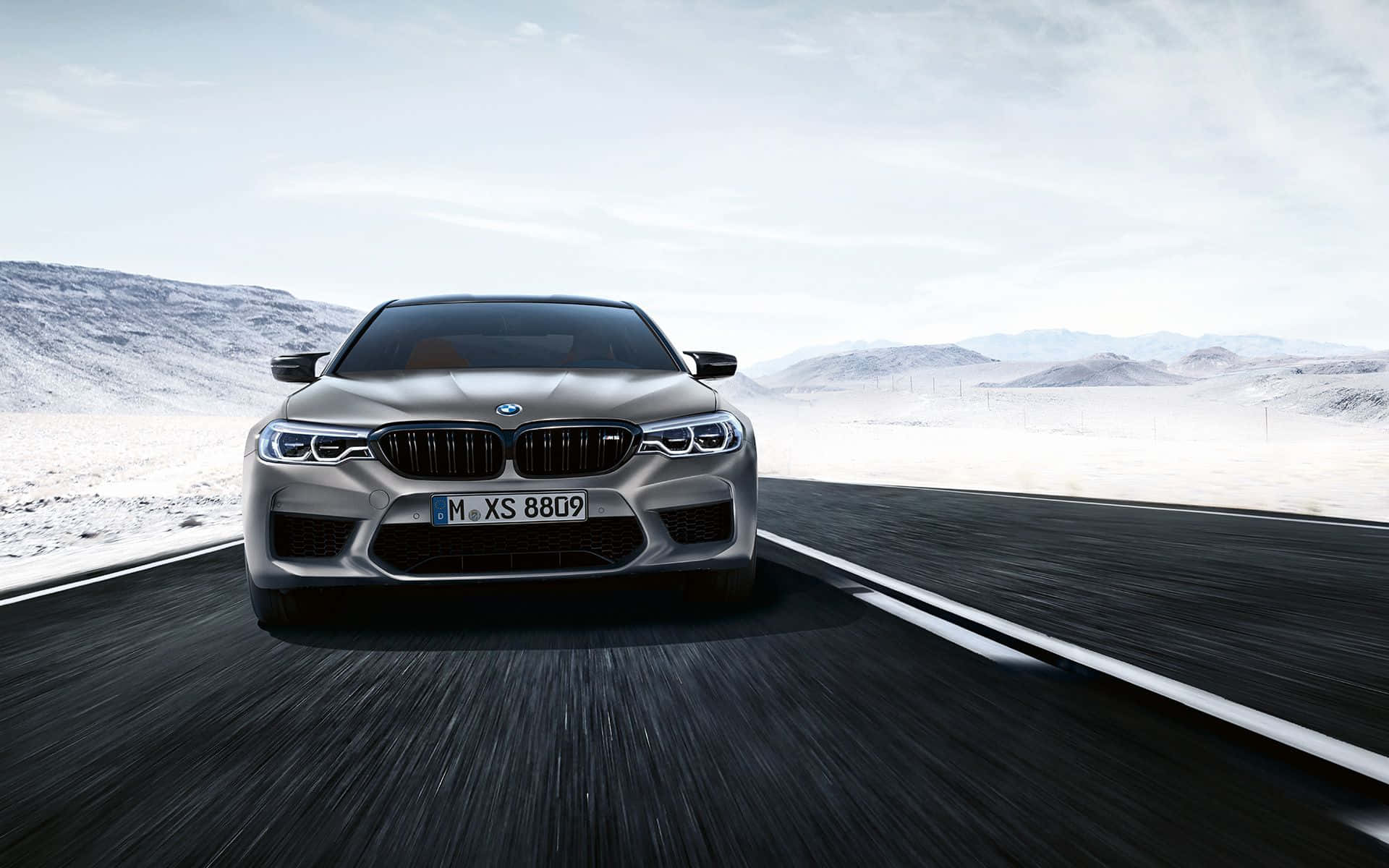 BMW 動圖 桌布 壁紙 — 一輛銀色BMW M5在明亮的天空下，沿著沙漠高速公路疾馳，遠處是積雪的山峰。