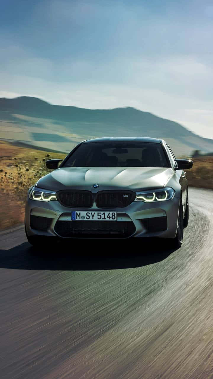 豪華車 桌布 壁紙 — 一輛銀色BMW M5在晴朗天空下，沿著蜿蜒的鄉村道路疾馳，背景是起伏的丘陵，帶有動態模糊效果。