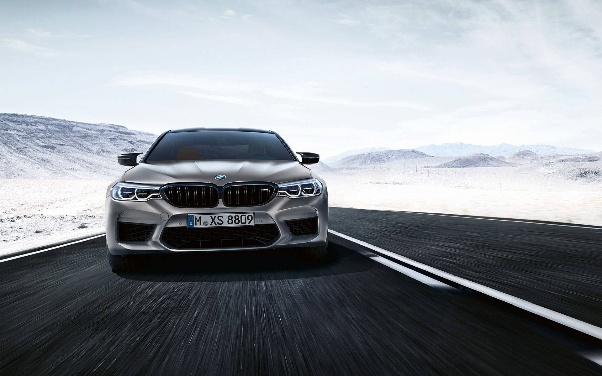 m 桌布 壁紙 — 一輛銀色BMW M5在沙漠高速公路上疾駛，遠方有白雪覆蓋的山脈，天空明亮。