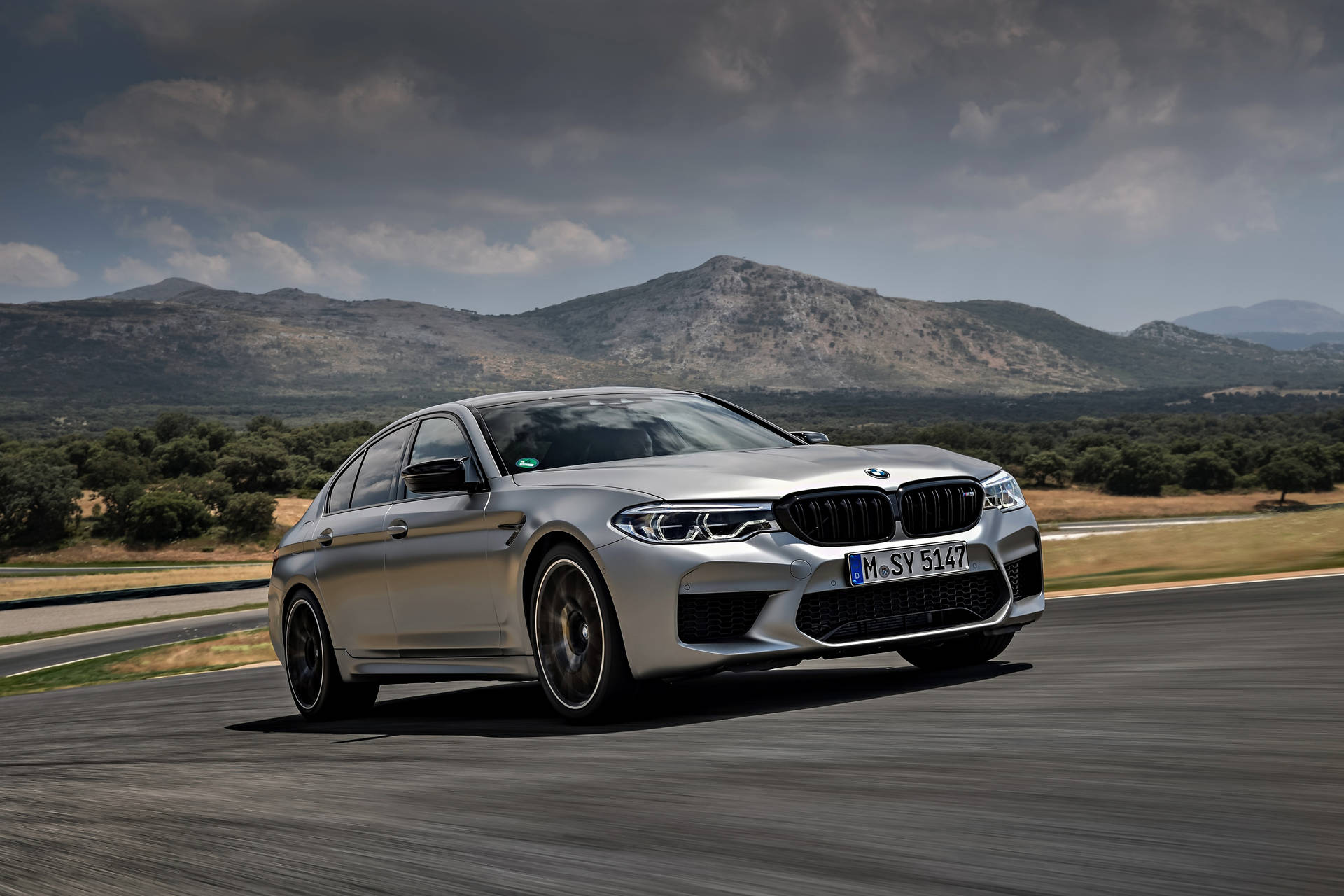 BMW 動圖 桌布 壁紙 — 一輛銀色BMW M5房車在戲劇性的多雲天空下沿著蜿蜒的山路飛馳。