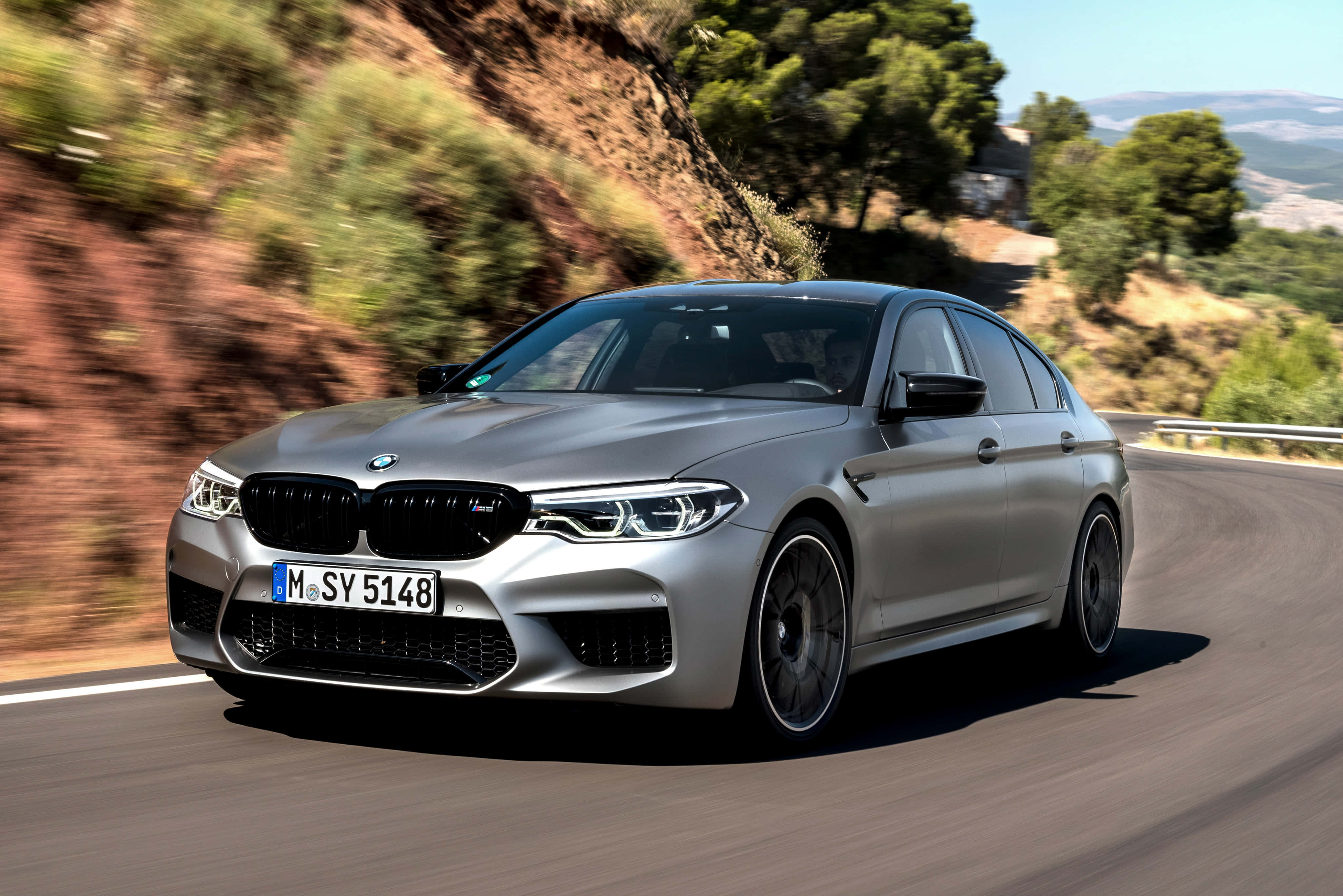 桌布 壁紙 — 一輛銀色BMW M5轎車在蜿蜒的山路上疾馳，背景是模糊的綠樹和岩石地形。