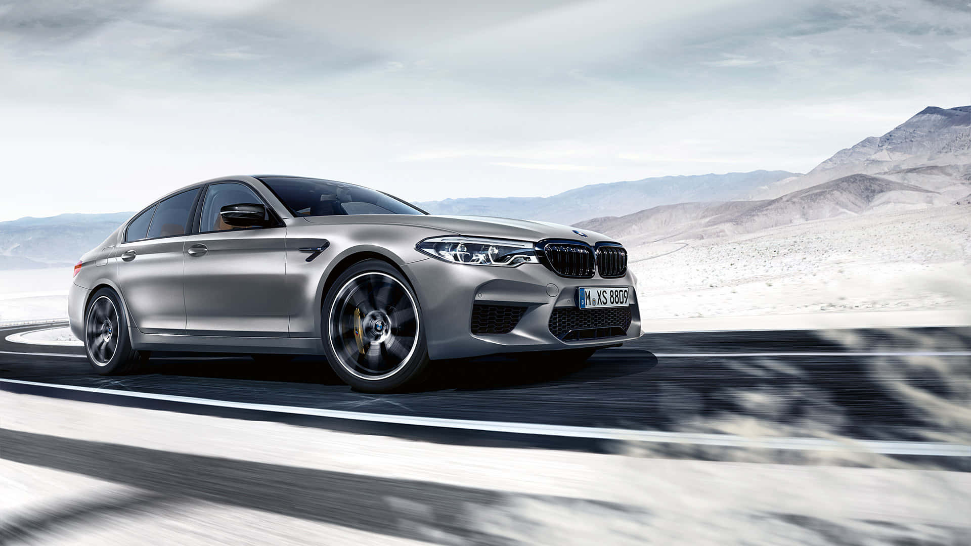桌布 壁紙 — 一輛銀色BMW M5轎車在陰天下沿著蜿蜒的沙漠公路疾駛，背景是山脈。