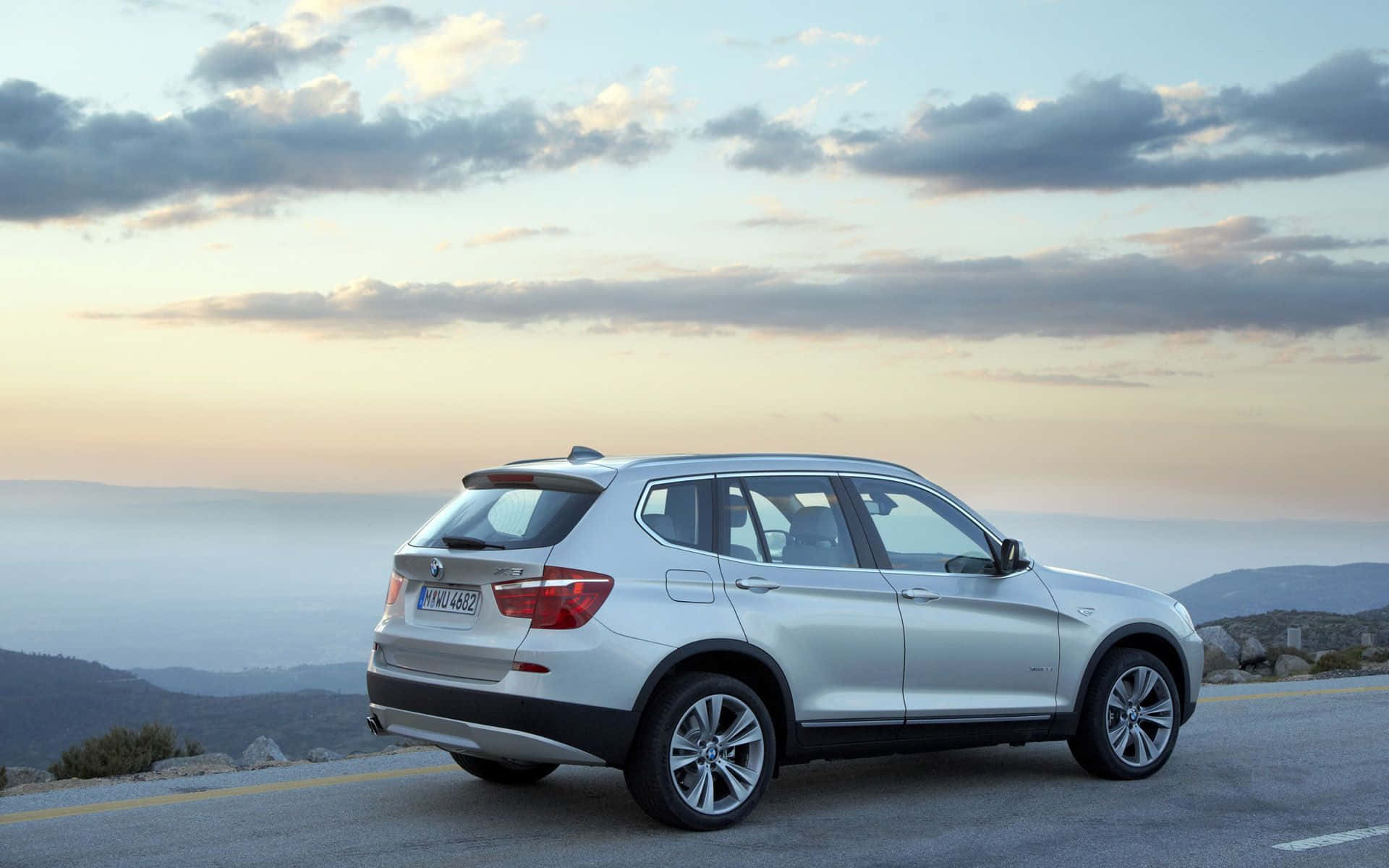 Bmw X3 桌布 壁紙 — 一輛銀色BMW SUV停泊在日落時分的海岸山路上，背景有戲劇性的雲層和遠山。