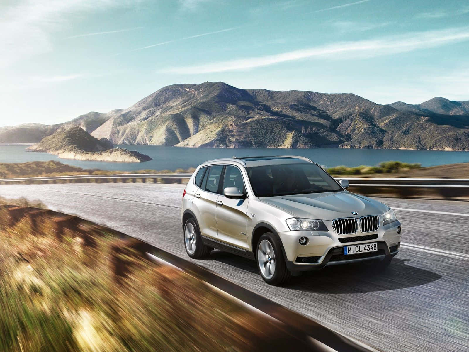 Bmw X3 桌布 壁紙 — 一輛銀色BMW SUV在晴朗藍天下沿著蜿蜒的海岸道路行駛，背景是山脈和湖泊。