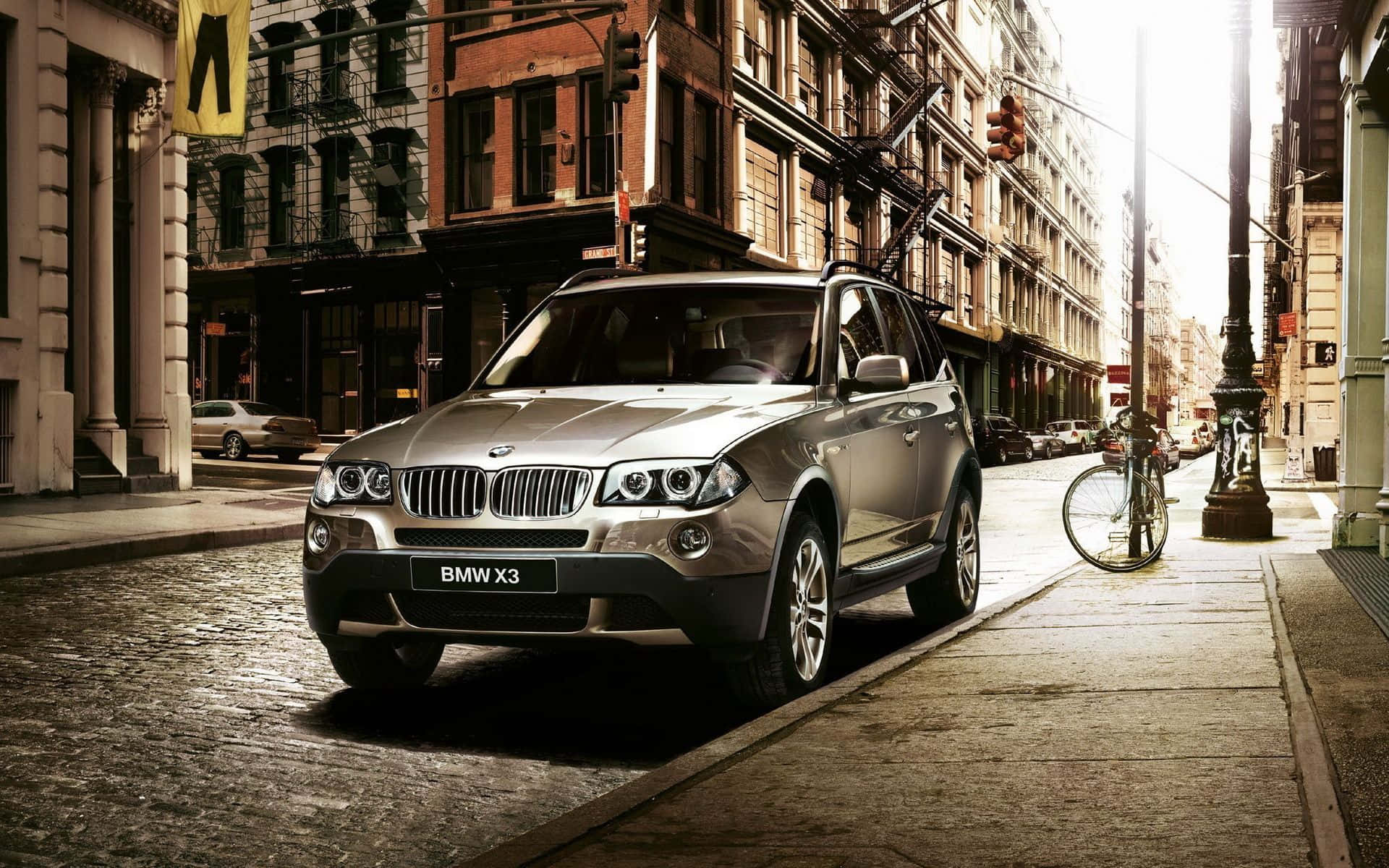 Bmw X3 桌布 壁紙 — 一輛銀色BMW X3 SUV停放在陽光灑落的鵝卵石街道上，兩旁是歷史悠久的磚造建築，附近有一輛腳踏車靠著。
