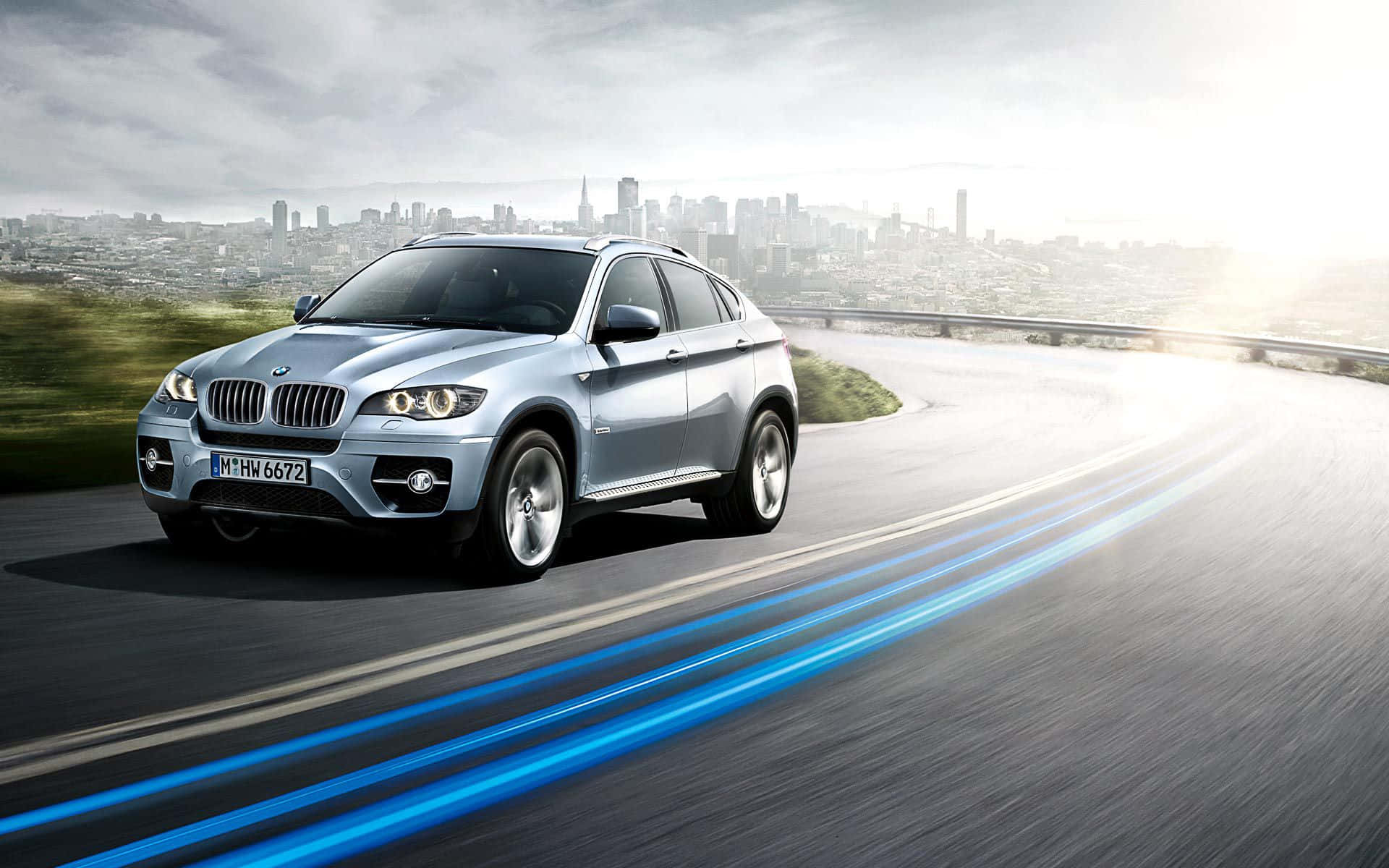 Bmw Laptop 桌布 壁紙 — 一輛銀色BMW X6 SUV在蜿蜒的高速公路上疾駛，遠處的城市天際線在薄霧中若隱若現。