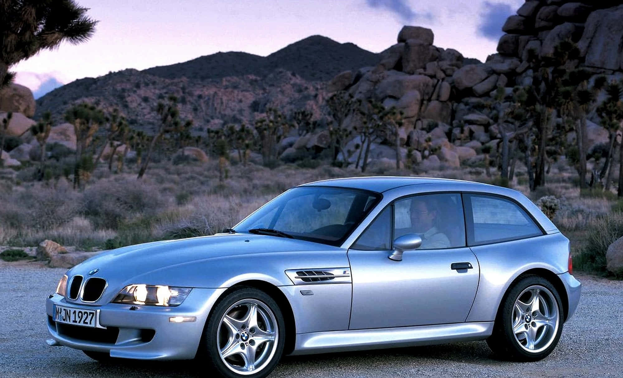 Bmw Z3 桌布 壁紙 — 一輛銀色BMW Z3敞篷跑車在暮色天空下閃閃發光，停泊在約書亞樹遍佈的岩石沙漠地帶的碎石路上。