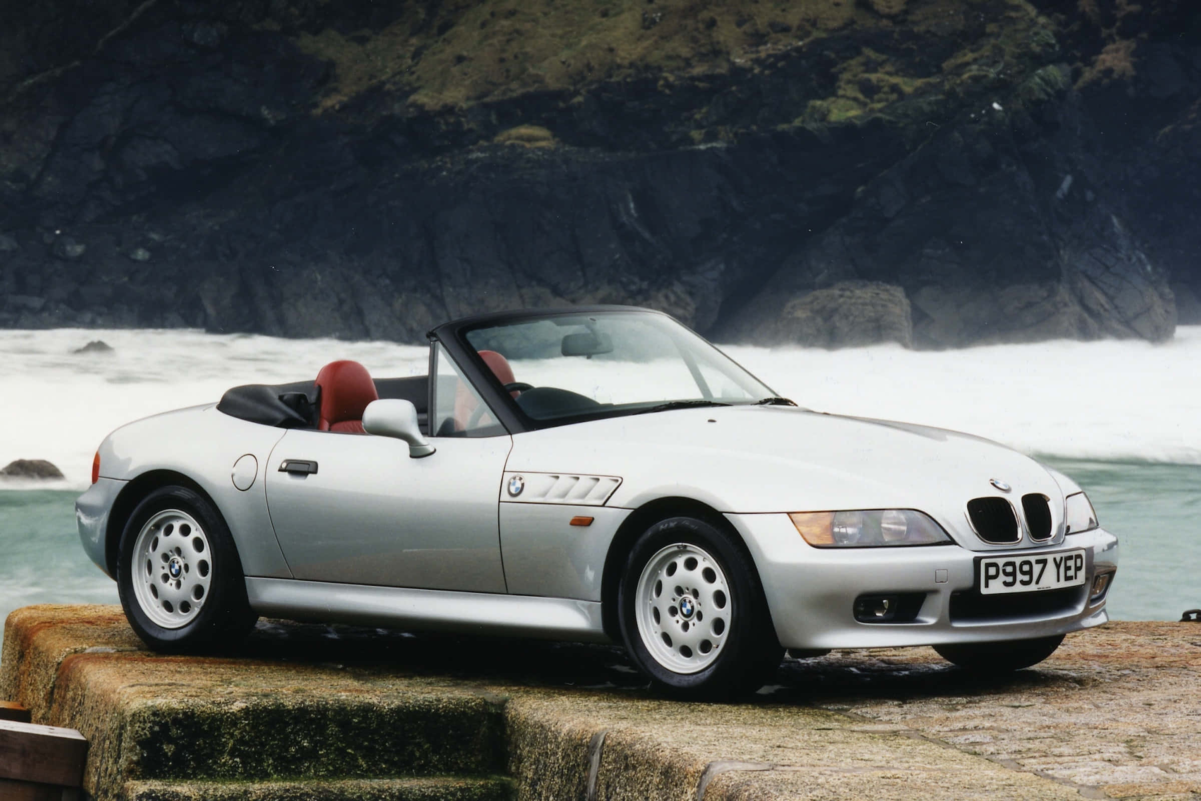 Bmw Z3 桌布 壁紙 — 一輛銀色BMW Z3敞篷跑車配紅色座椅，停泊在石頭上，旁邊是拍打暗黑懸崖的洶湧海浪。