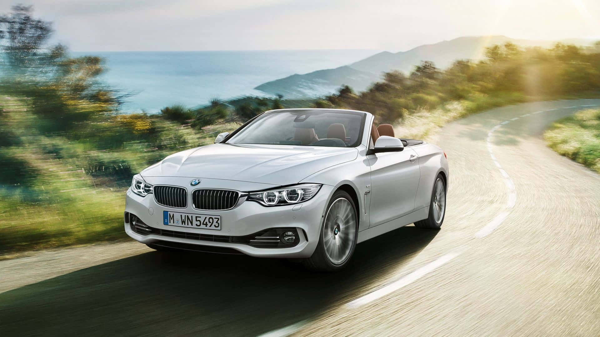 Bmw 4 Series 桌布 壁紙 — 一輛銀色BMW敞篷車在明亮陽光下，沿著海岸公路奔馳，背景是模糊的綠樹與海景。