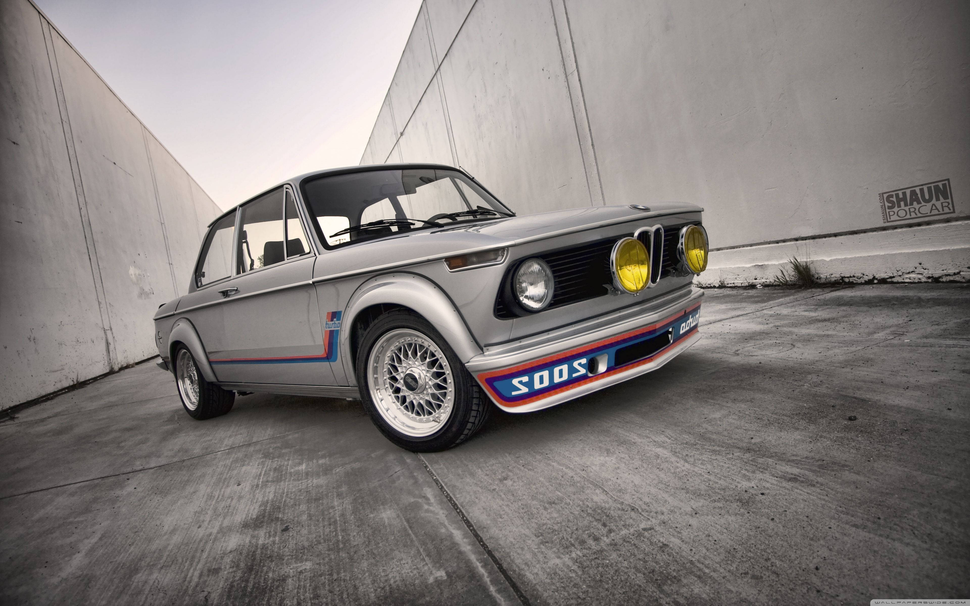 BMW 動圖 桌布 壁紙 — 一輛銀色BMW賽車，帶有黃色頭燈和藍紅條紋，停在混凝土隧道中。