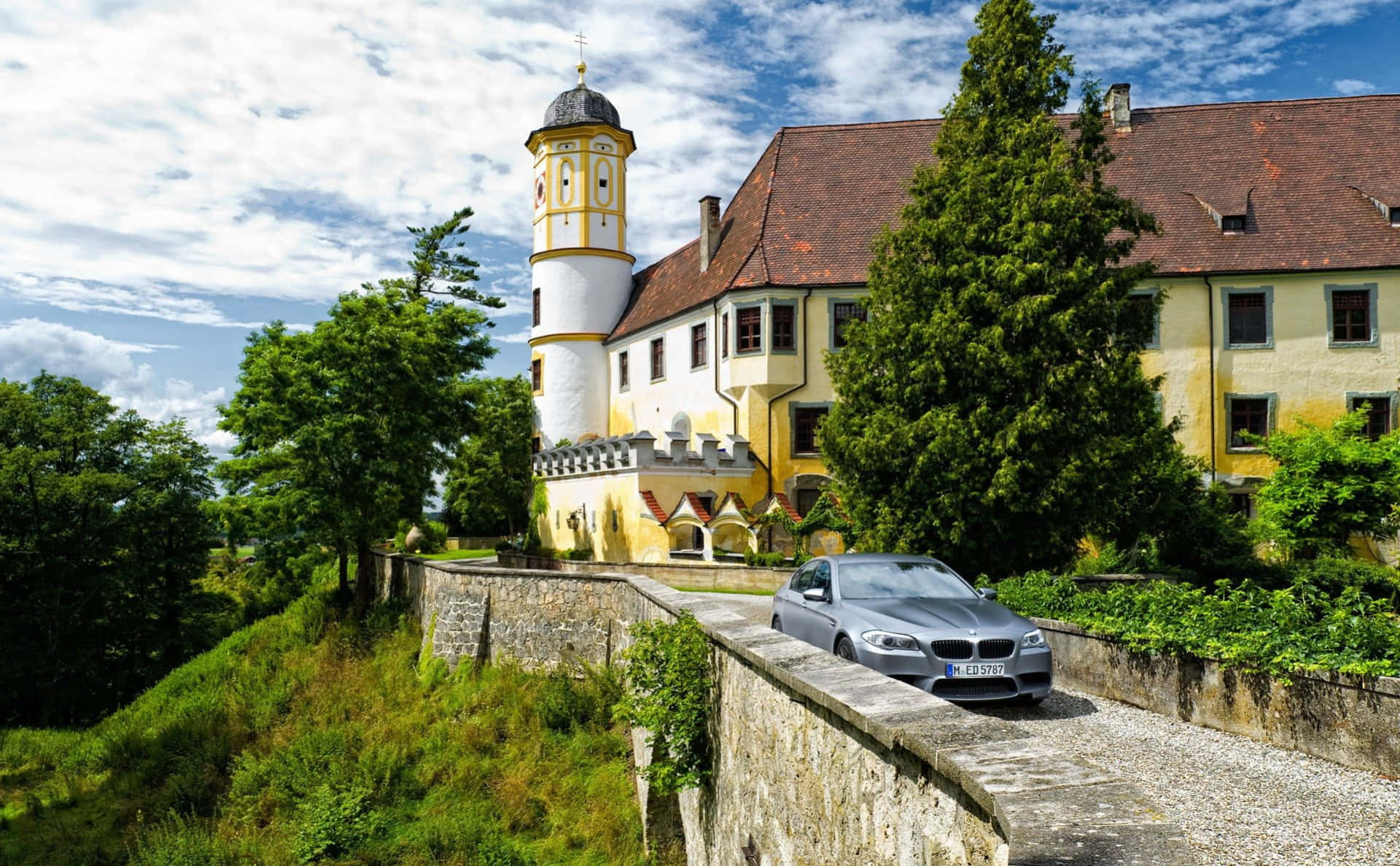 Driveway Ideas 桌布 壁紙 — 一輛銀色BMW轎車沿著石徑行駛，旁邊是一座歷史悠久的黃白色城堡，城堡有塔樓，天空部分多雲。