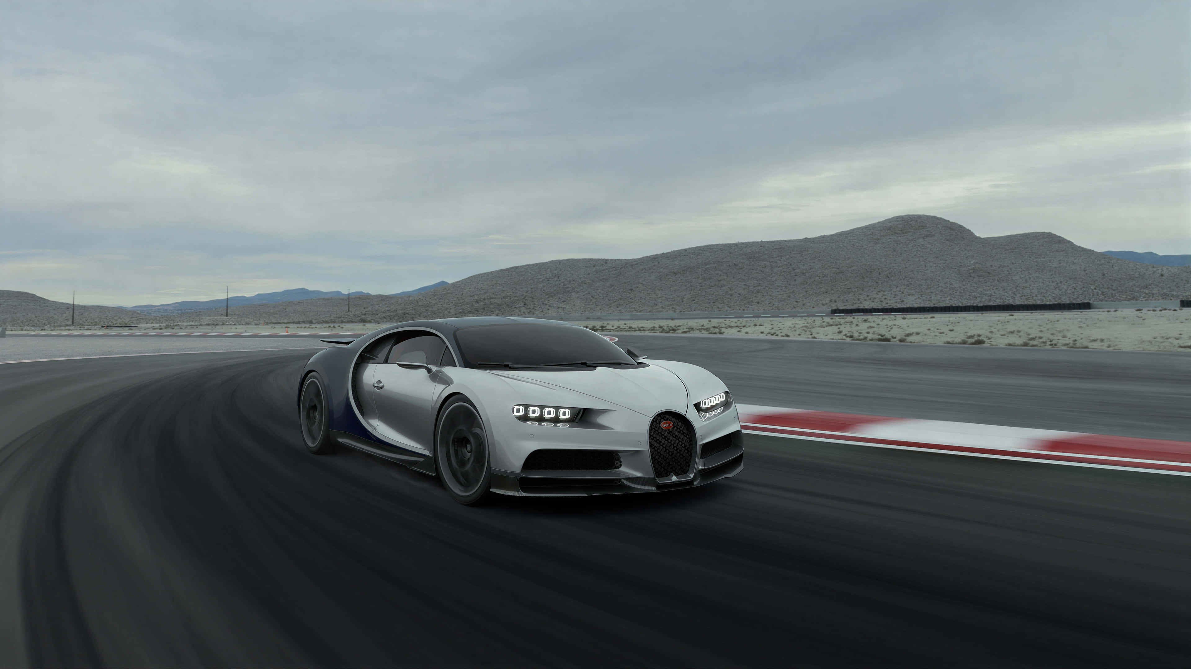 bugatti iPhone 桌布 壁紙 — 一輛銀色Bugatti Chiron在沙漠賽道上疾馳，帶有模糊的動態效果，遠方是岩石丘陵，天空多雲。