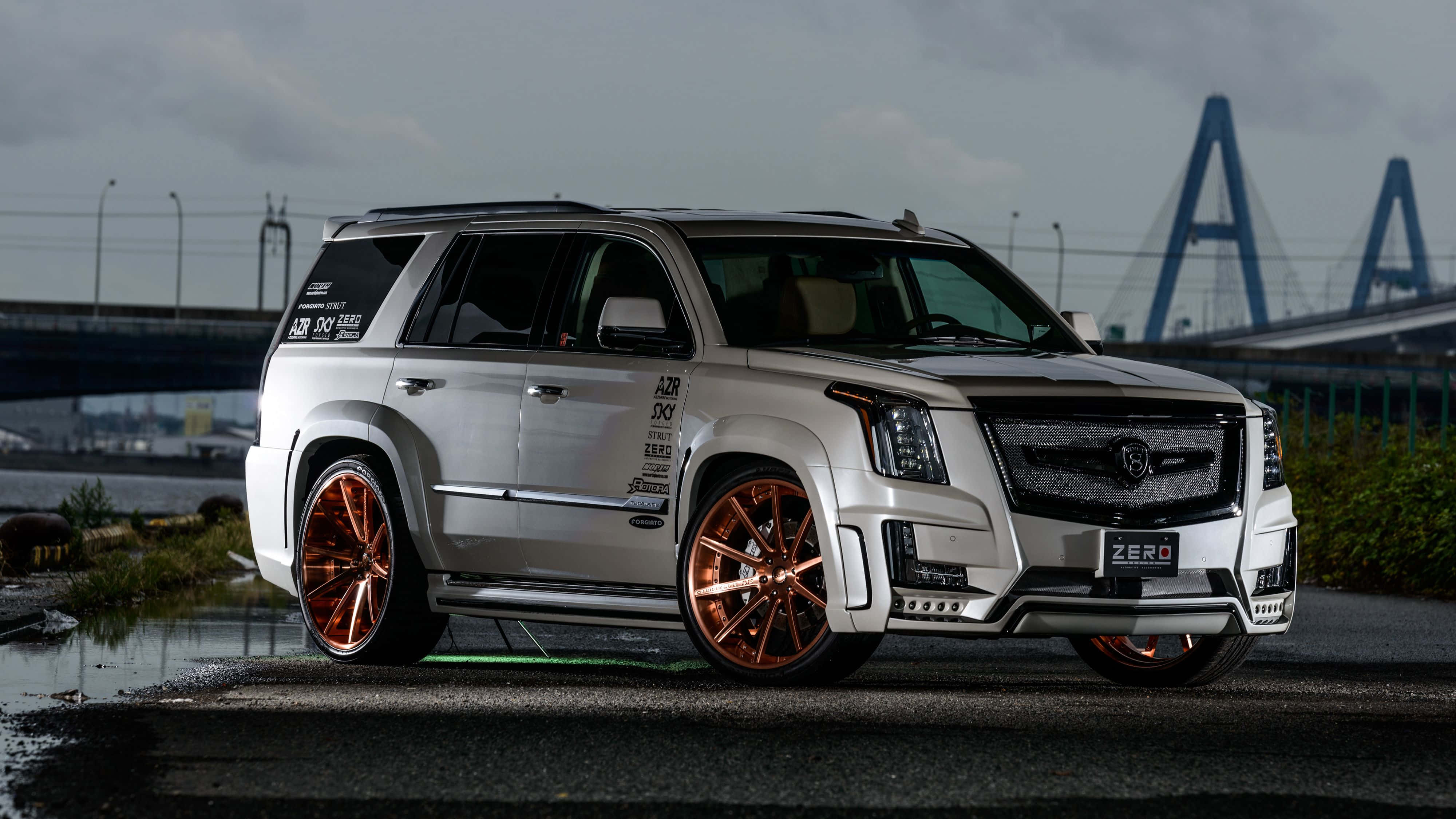Cadillac Escalade 桌布 壁紙 — 一輛銀色Cadillac SUV配備銅色輪圈，停放在濕漉漉的柏油路上，背景是陰天和一座藍色橋樑。