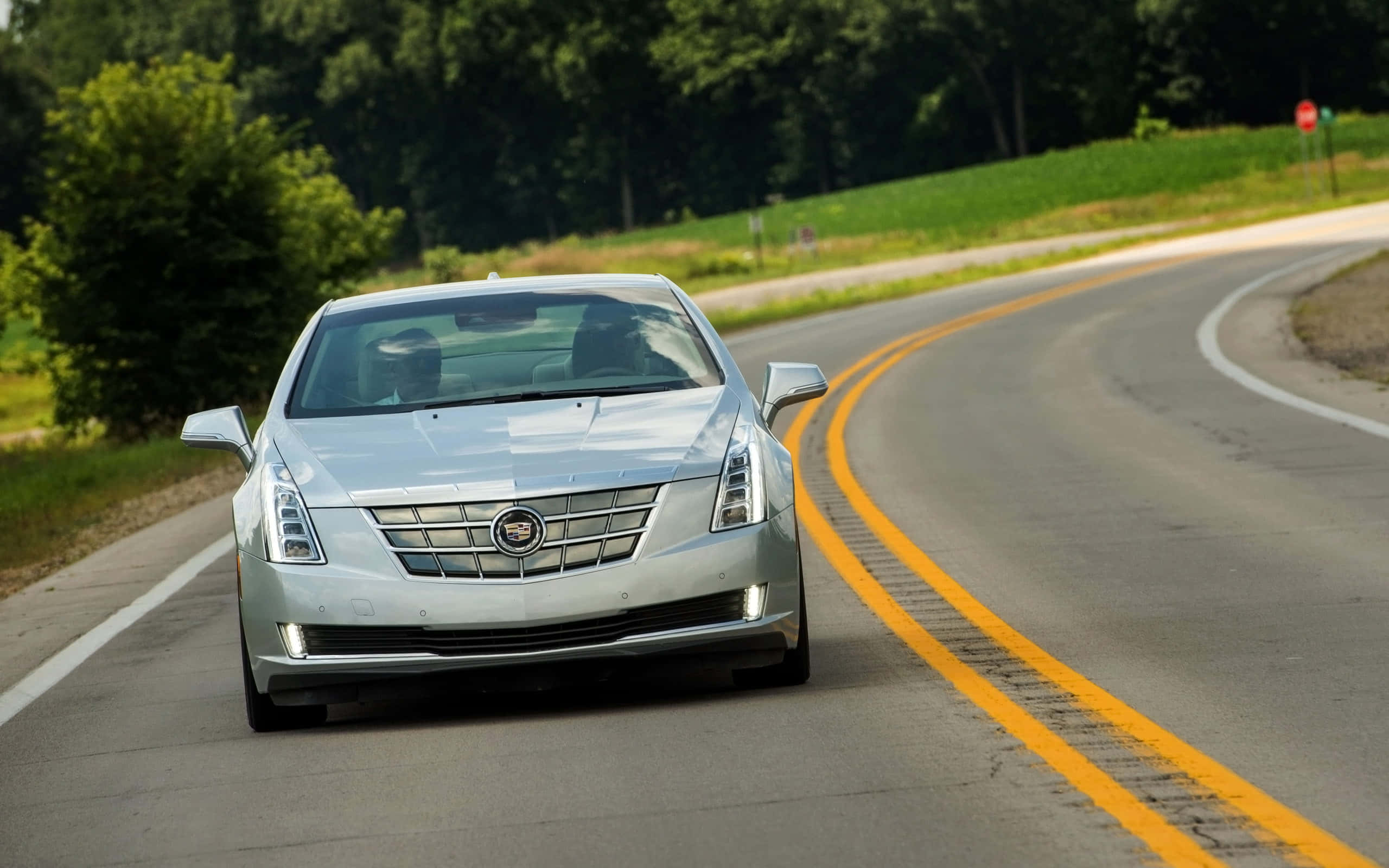 Cadillac Elr 桌布 壁紙 — 一輛銀色Cadillac轎車在明亮的日光下，沿著一條蜿蜒的鄉村道路駛向鏡頭，道路兩旁綠樹成蔭，草地青翠。