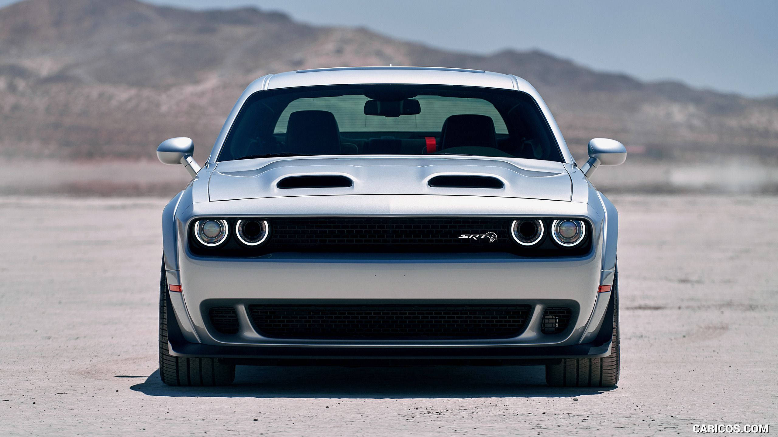 Challenger 桌布 壁紙 — 一輛銀色Dodge Challenger SRT Hellcat在塵土飛揚的沙漠平原上正面朝前，背景是崎嶇的山脈，天空晴朗。