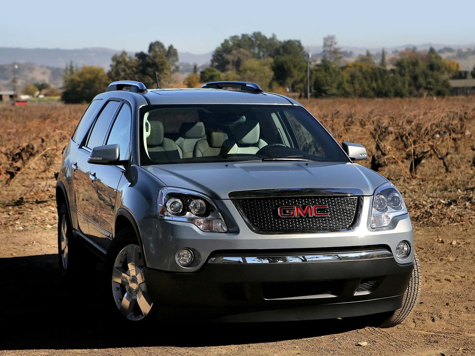 Gmc Acadia 桌布 壁紙 — 一輛銀色GMC Acadia SUV停放在泥土路上，周圍是乾燥的葡萄園行和遠方的山丘，天空清澈湛藍。