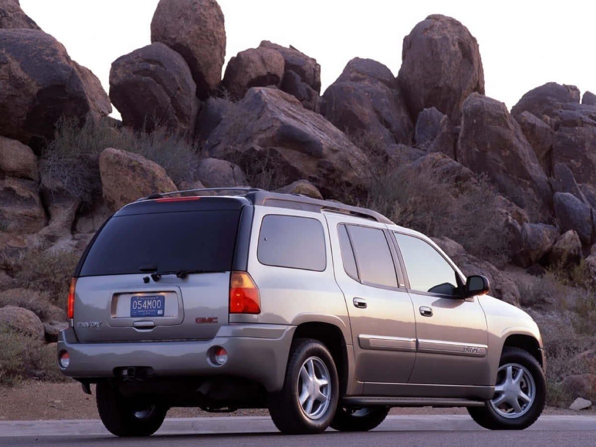 Gmc Envoy 桌布 壁紙 — 一輛銀色GMC Envoy SUV停放在鋪砌的道路上，背景是沙漠，有巨大的粗糙岩石和乾燥的灌木。