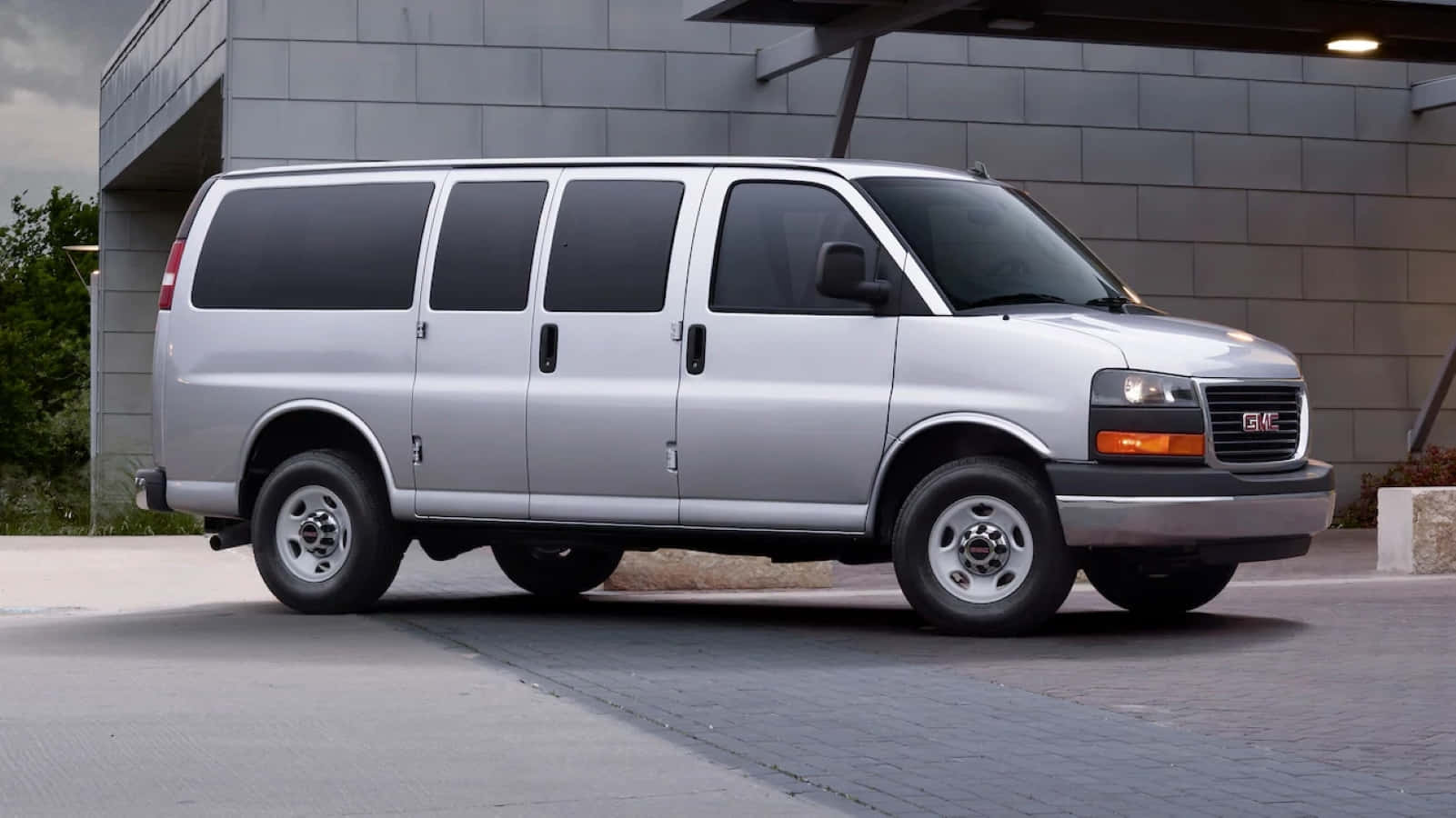 Gmc Savana 桌布 壁紙 — 一輛銀色GMC客貨車停放在鋪砌的停車場上，旁邊是一座現代灰色磚砌建築，天空多雲。