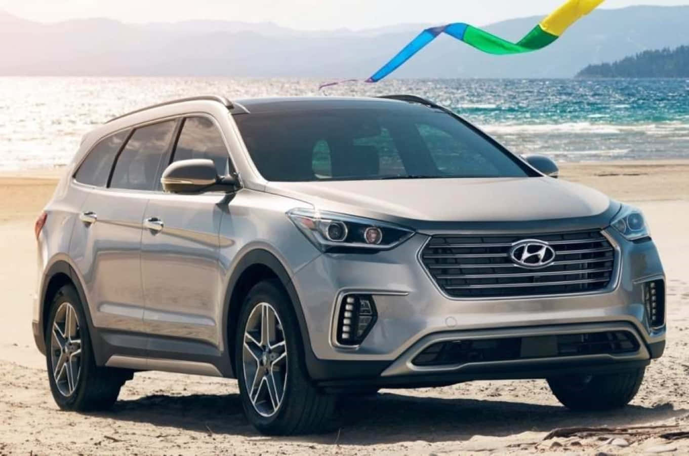 Hyundai Veracruz 桌布 壁紙 — 一輛銀色Hyundai SUV停泊在陽光明媚的海灘上，海面上波濤洶湧，頭頂上飛著色彩繽紛的風箏。