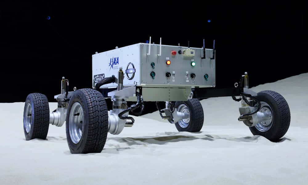 Lunar Rover 桌布 壁紙 — 一輛銀色JAXA月球車配備堅固輪胎，站在模擬月球表面，天空黑暗，遠處有藍色燈光。