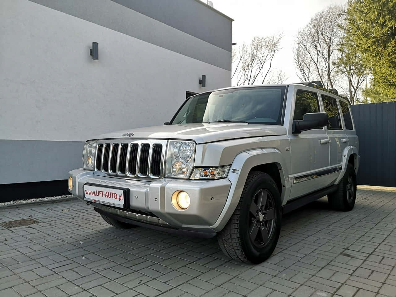 Jeep Commander 桌布 壁紙 — 一輛銀色Jeep Commander SUV停放在灰色磚砌車道上，旁邊是一座現代建築，背景有光禿禿的樹木。