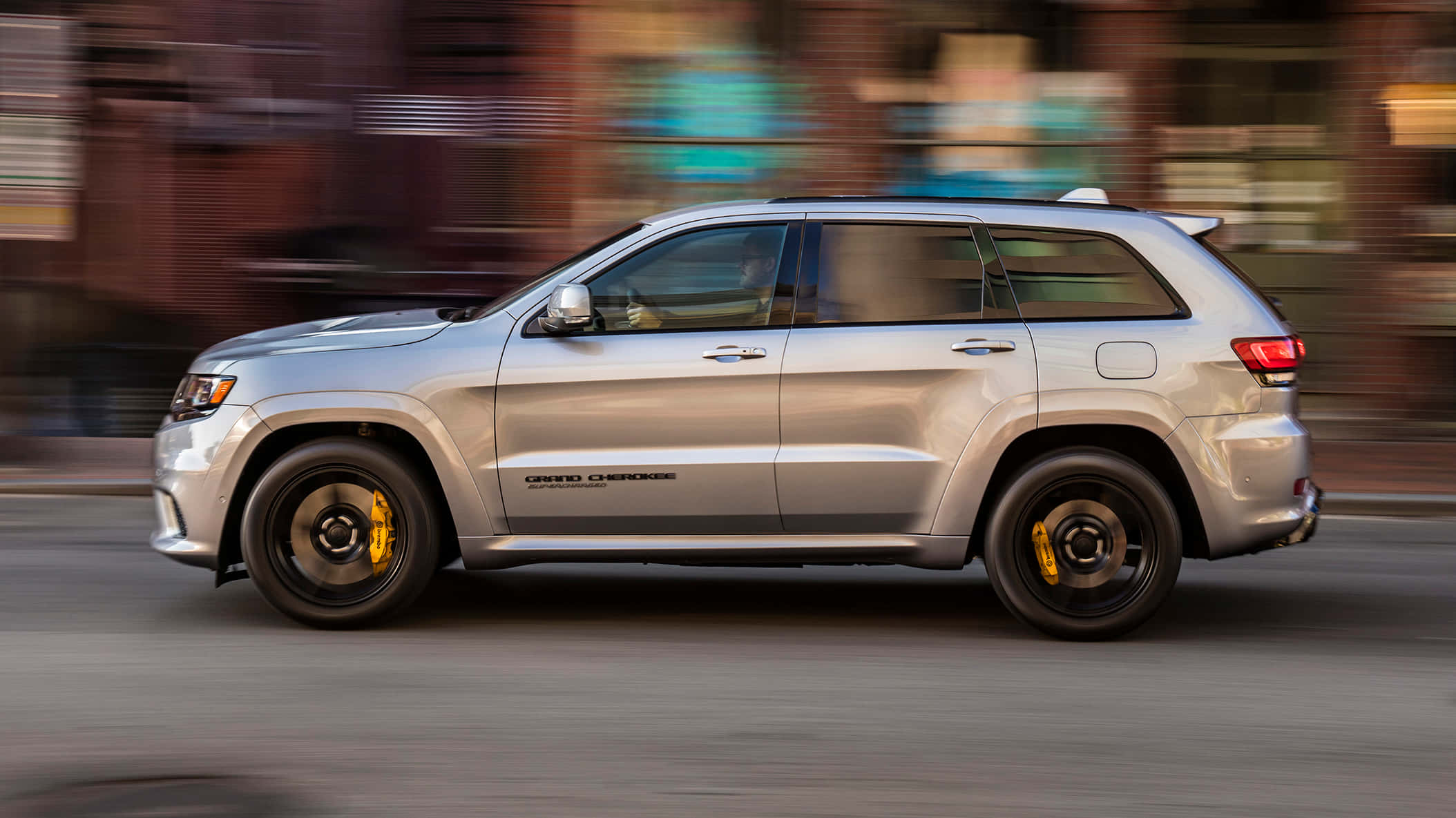 Jeep Trackhawk 桌布 壁紙 — 一輛銀色Jeep Grand Cherokee在模糊的磚砌建築背景下於城市街道上飛馳。