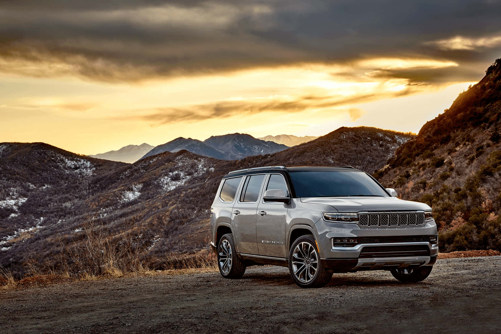 Jeep Wagoneer 桌布 壁紙 — 一輛銀色Jeep Grand Wagoneer在日落時分停泊在泥土路上，背景是積雪的山脈。