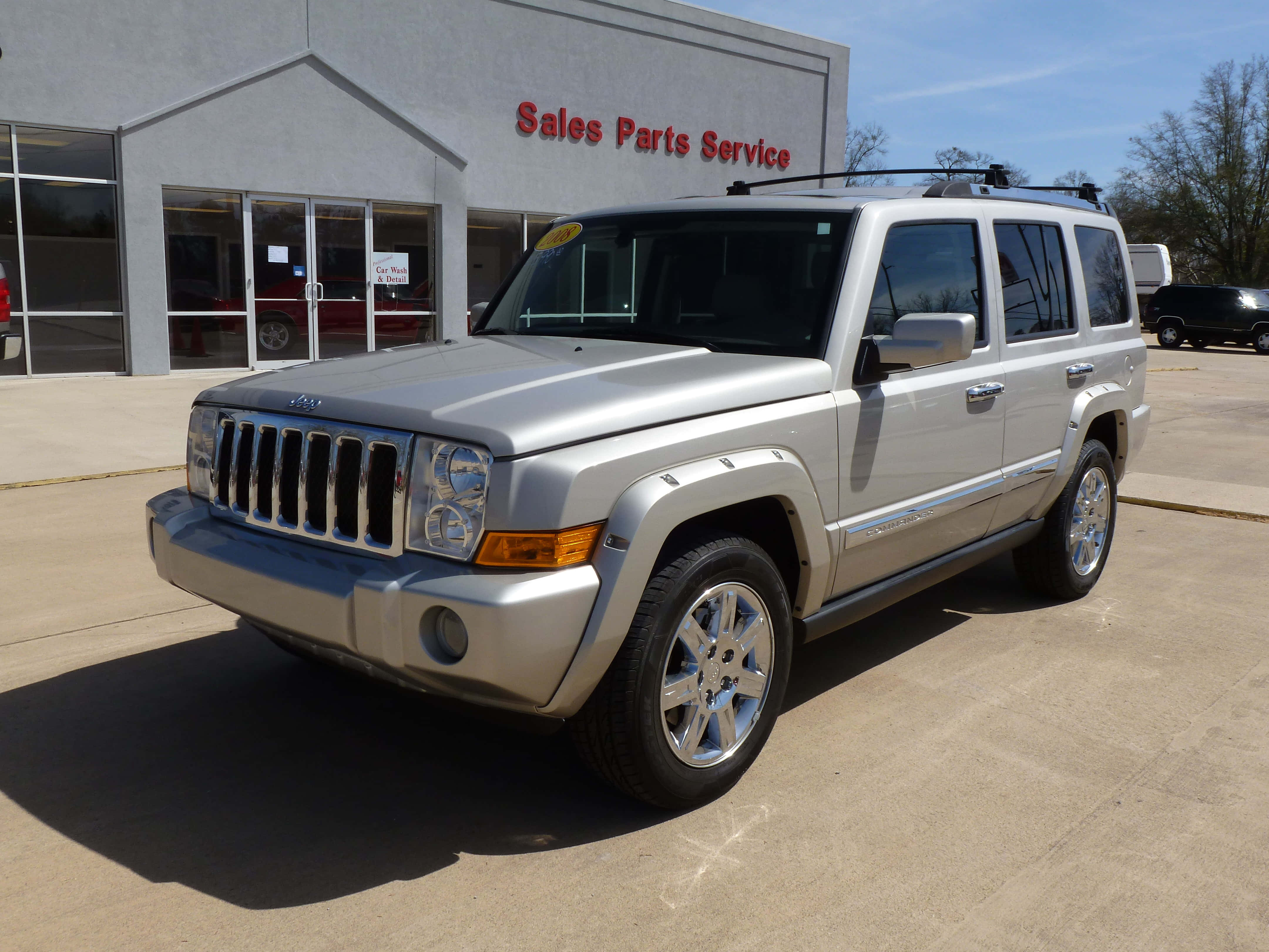Jeep Commander 桌布 壁紙 — 一輛銀色Jeep Liberty SUV停放在汽車經銷商前，背景是晴朗的藍天和樹木。