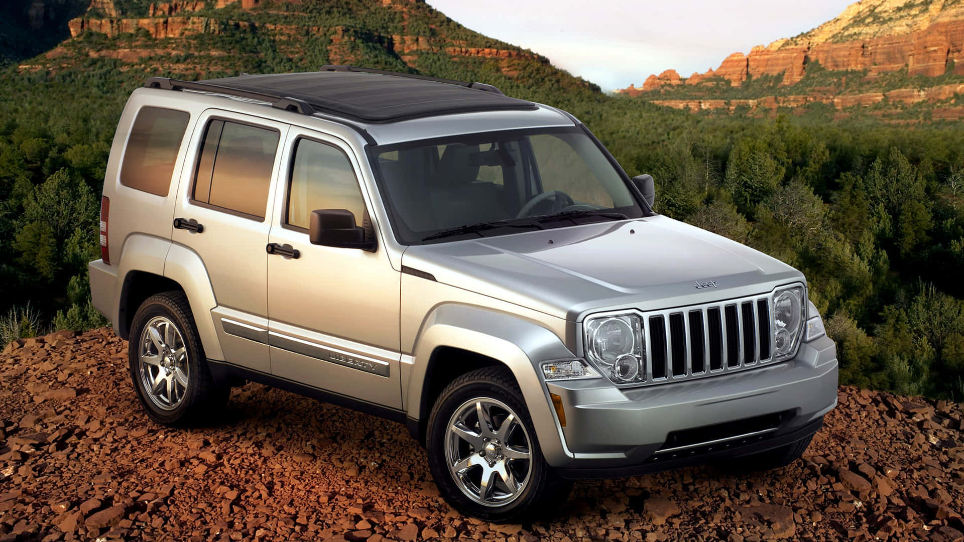 Jeep Liberty 桌布 壁紙 — 一輛銀色Jeep Liberty SUV停放在岩石地形上，背景有紅色岩層和綠色樹木，天空柔和。
