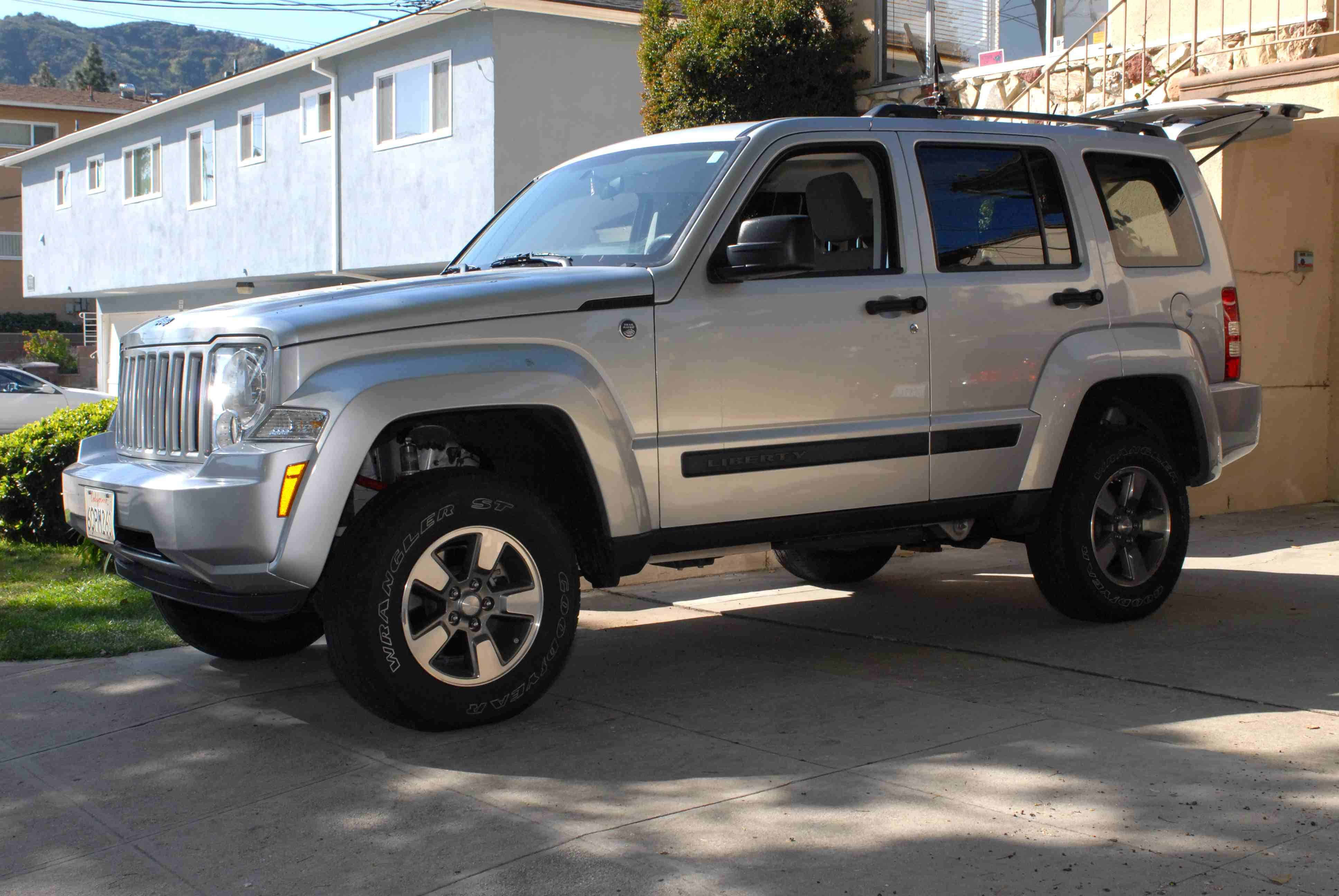 Jeep Liberty 桌布 壁紙 — 一輛銀色Jeep Liberty SUV停放在車道上，背景是藍色房屋和綠色灌木叢，在明亮的陽光下。