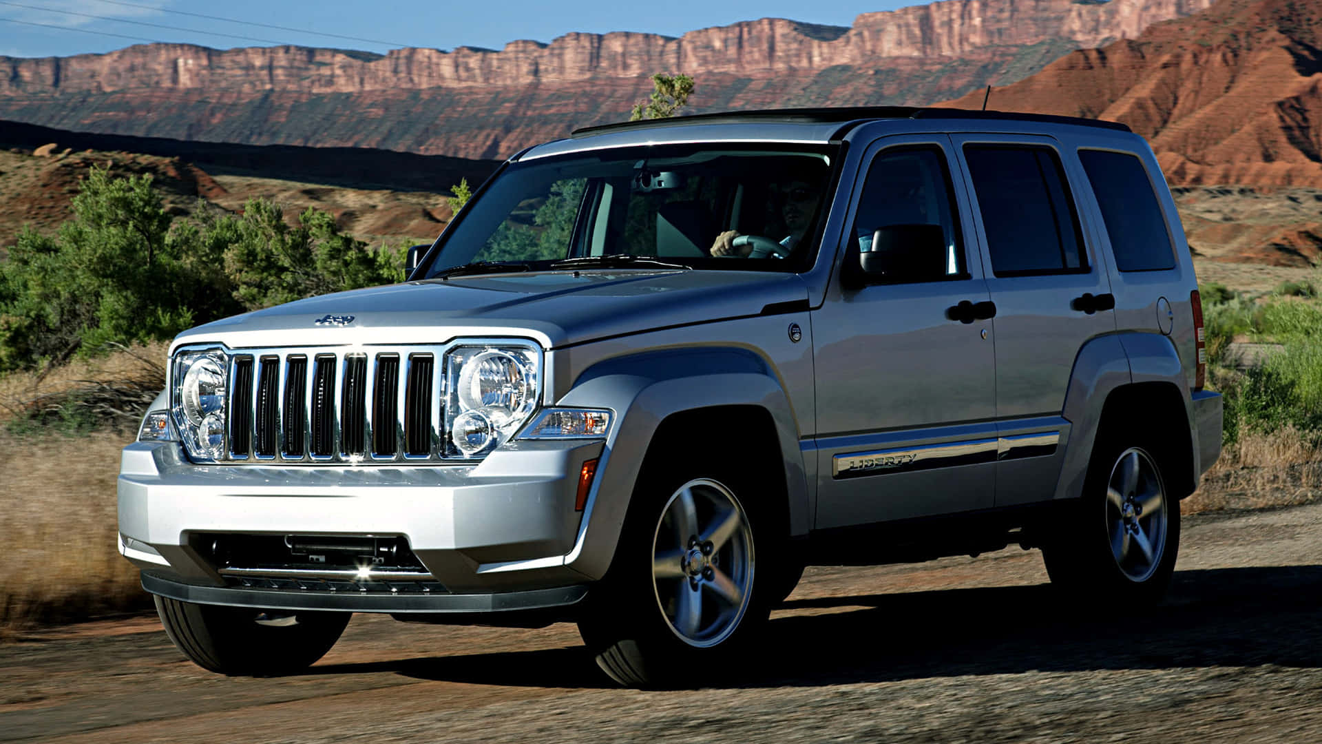 Jeep Liberty 桌布 壁紙 — 一輛銀色Jeep Liberty SUV停泊在泥土路上，背景是紅色岩石峽谷懸崖，天空清澈湛藍。