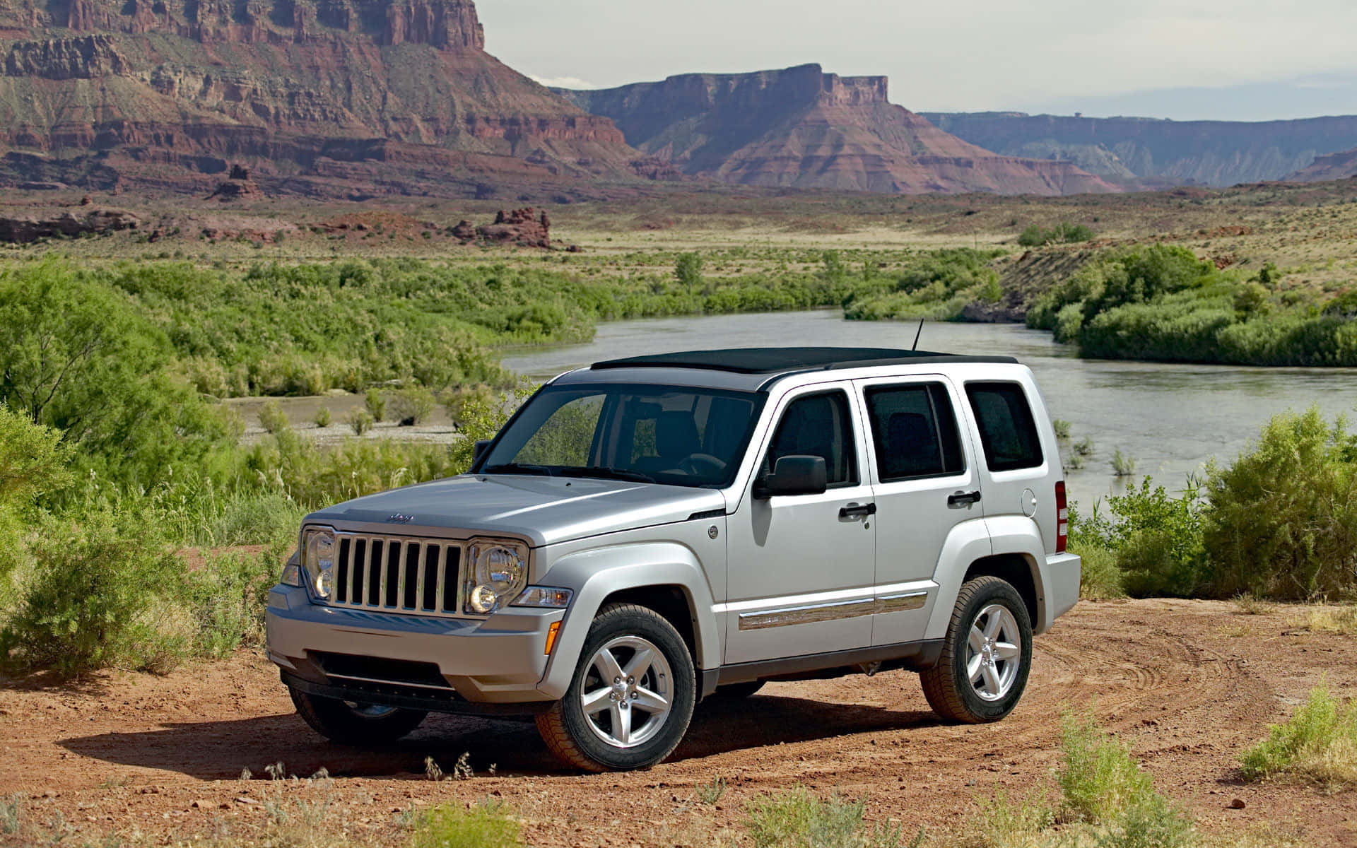 Jeep Liberty 桌布 壁紙 — 一輛銀色Jeep Liberty停泊在河邊的泥路上，背景是紅色岩石峽谷懸崖。