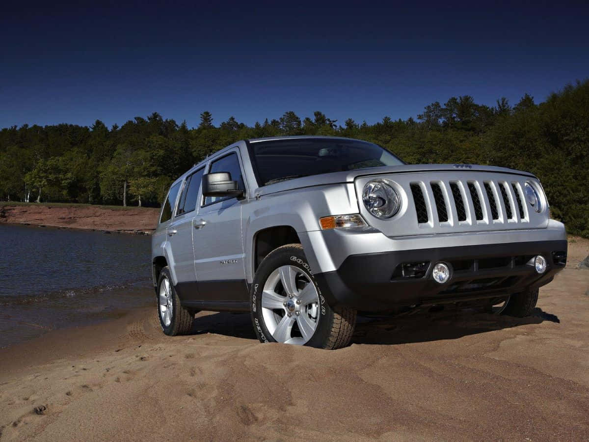 Jeep Patriot 桌布 壁紙 — 一輛銀色Jeep Patriot SUV停泊在沙質岸邊，旁邊是平靜的水面，背後有茂密森林，天空清澈湛藍。