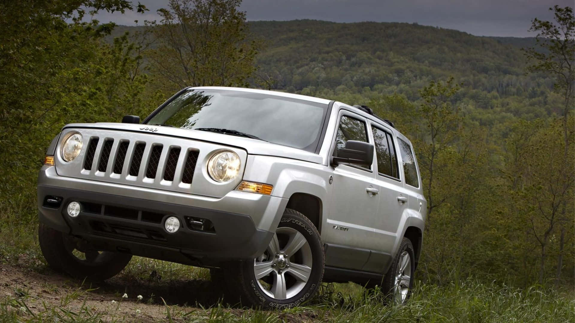 Jeep Patriot 桌布 壁紙 — 一輛銀色Jeep Patriot SUV停放在草地小徑上，周圍是茂密的綠色森林和起伏的丘陵，天空多雲。