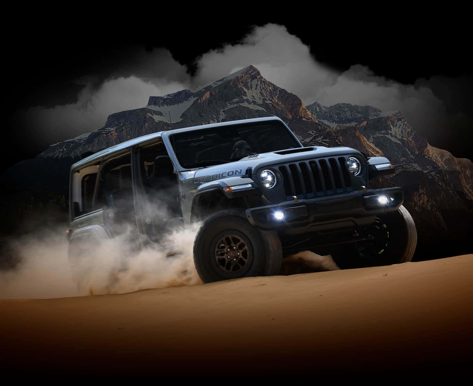 吉普 桌布 壁紙 — 一輛銀色Jeep Wrangler Rubicon在夜間駛過沙漠，揚起沙塵，背景是白雪覆蓋的山脈，天空烏雲密佈。