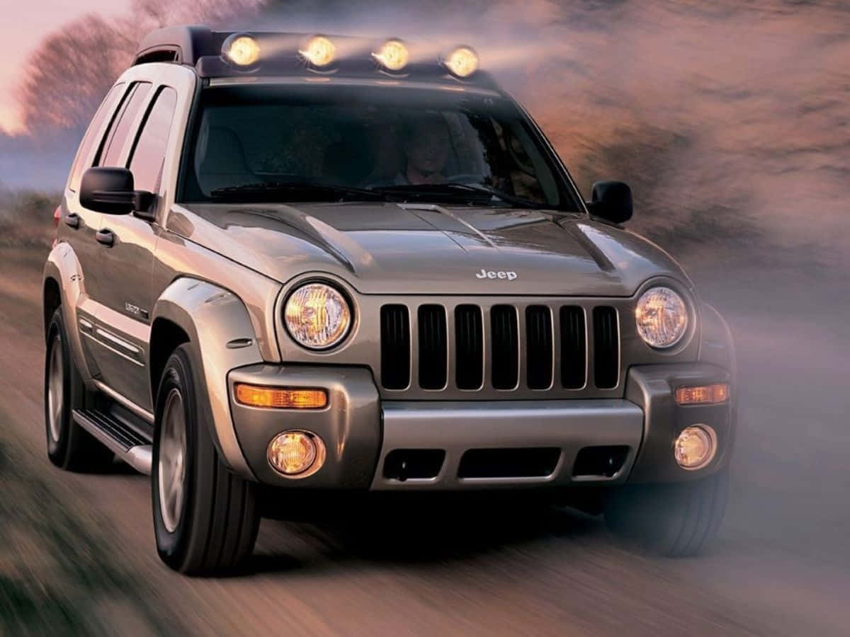 Jeep Liberty 桌布 壁紙 — 一輛銀色Jeep Wrangler在塵土飛揚的道路上疾馳，車頭燈在朦朧的日落背景下閃耀。