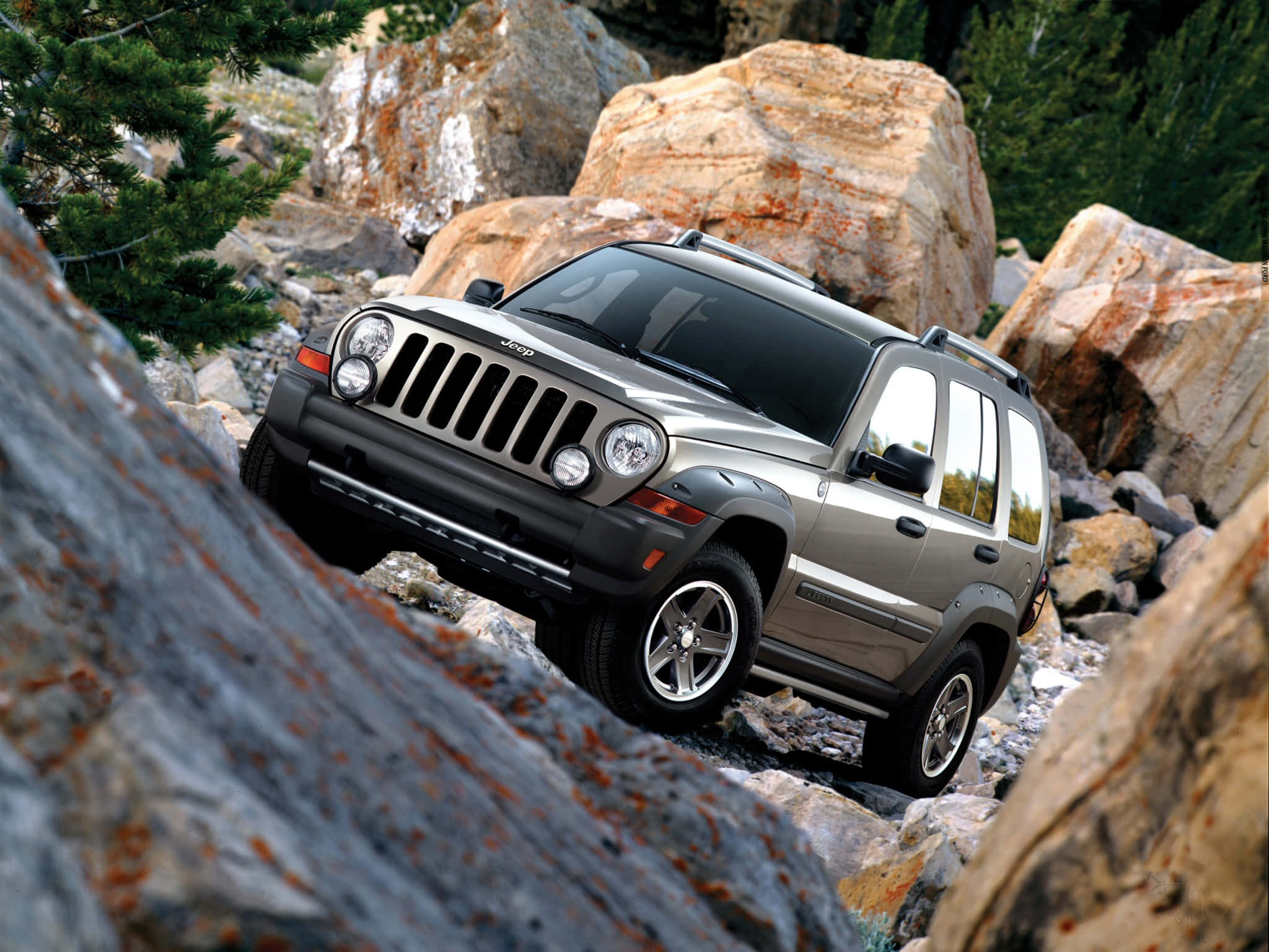 Jeep Liberty 桌布 壁紙 — 一輛銀色Jeep Wrangler在松樹和巨石環繞的崎嶇岩石地形上行駛。