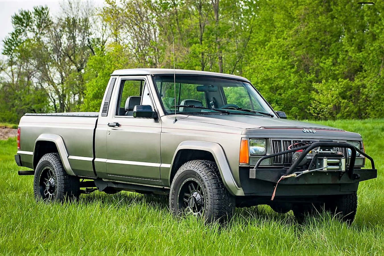 Jeep Comanche 桌布 壁紙 — 一輛銀色Jeep皮卡車，配備黑色車輪和前保險桿絞盤，停在高高的綠色草地上，背景有樹木。